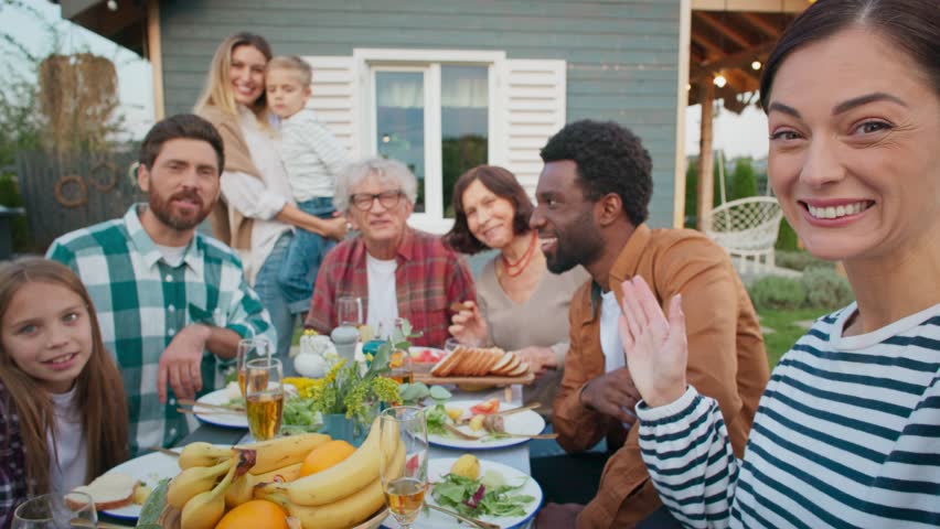 Group of people sitting together at table outdoors. Family gathering in village. Visiting parents. Looking at camera while waving. Celebrating something important. Enjoying tasty meals.