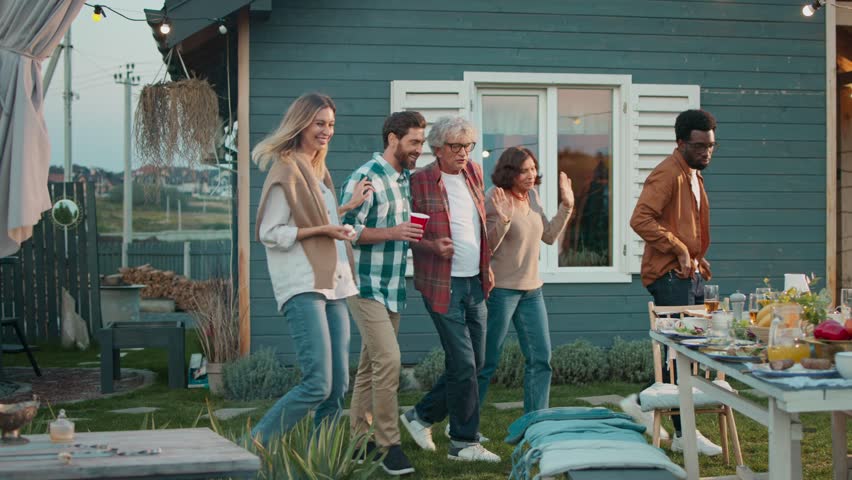 Warm moment of family coming to table placed on green lawn. Everyone sitting down. Getting ready for eating tasty meal. Table covered with many dishes. Kids running to parents. Dinner.