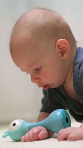 baby in a blue lies on their tummy on soft play mat, smiling while reaching toy, infant is learning to crawl, developing motor skills and coordination, home playtime, baby growth, vertical