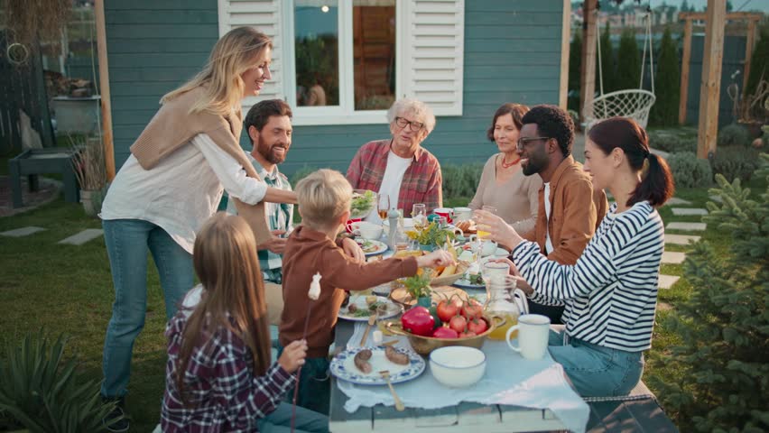 People gathering together at table. Woman bringing dish and placing to other plates. Family thanking woman about tasty food. Female blushing. Covering face with both hands. Picknick.