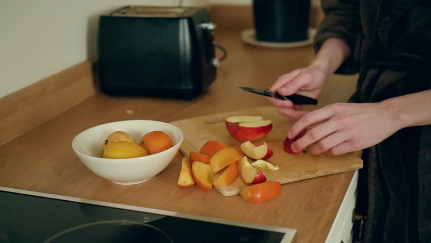 Man slicing apple and persimmon on wooden board in kitchen. Male cutting fresh fruits into small pieces. Guy preparing healthy snack with mixed produce. Person chopping natural ingredients for meal