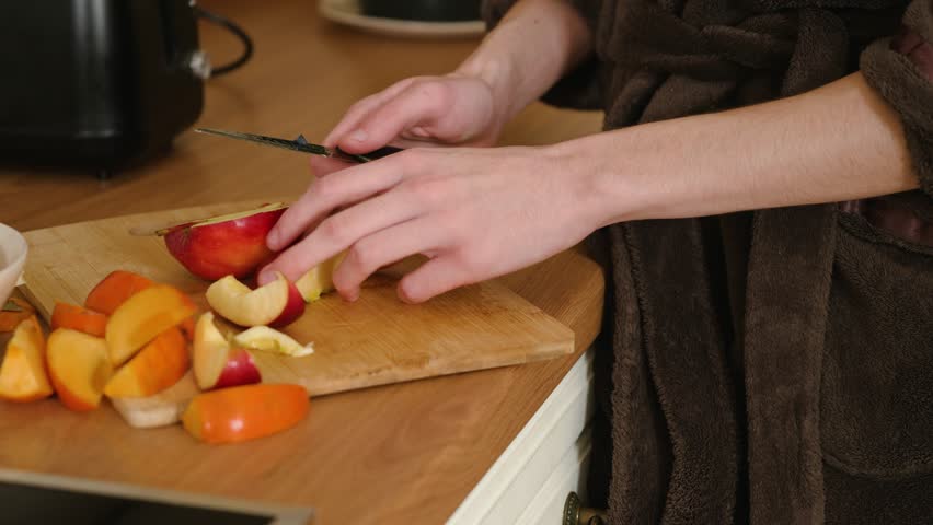 Man slicing apple on wooden cutting board in kitchen. Male carefully cutting fruit into smaller pieces with knife. Guy preparing fresh apple slices for snack. Person handling ripe produce while making