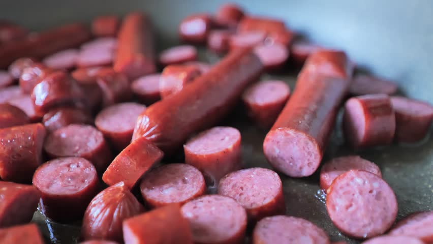 Close-up of sliced and whole sausages frying in a pan, glistening with oil - 4K