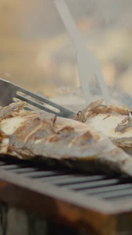 Vertical Screen: Succulent sea bream fish grilling on barbecue, chef carefully turning seafood with metal spatula over sizzling flames