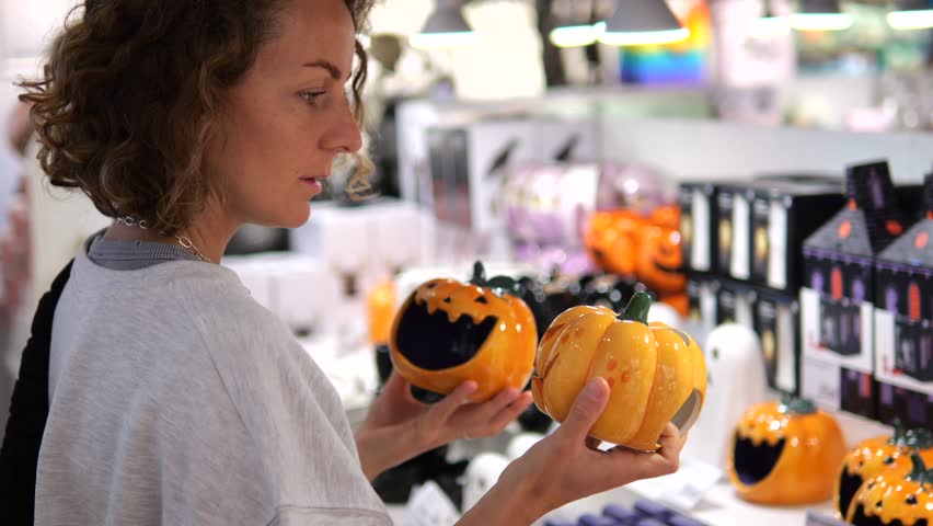 Young woman browsing seasonal decorations, comparing small orange ceramic pumpkins while standing in supermarket aisle during festive halloween shopping experience