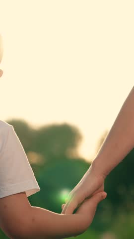 Happy kid walks through park with green trees holding her mom hand and smiling. Mom son, holding hand, walking in park on summer day. Concept family relations. Happy family relaxing together in nature