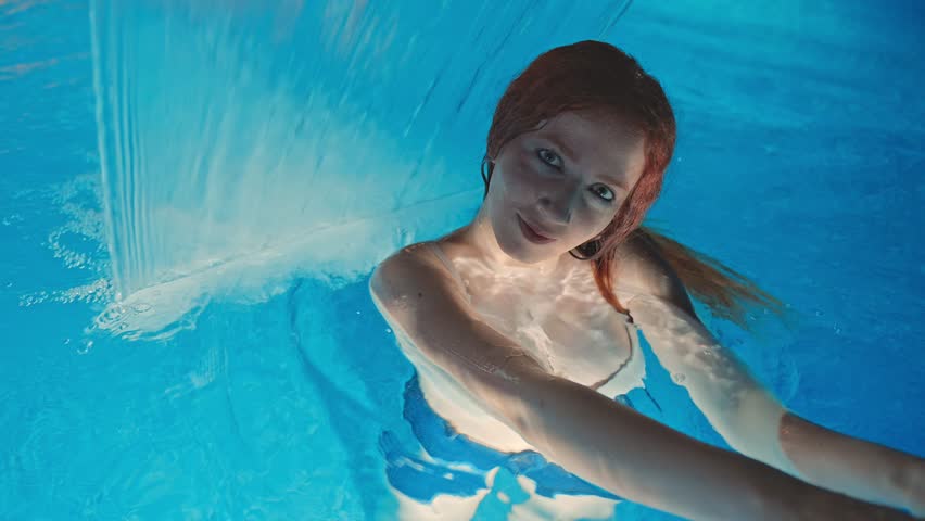 A young red-haired woman smiling in a brightly lit swimming pool at night. The reflections of the lights create a relaxing and summery atmosphere, evoking vacation and leisure.