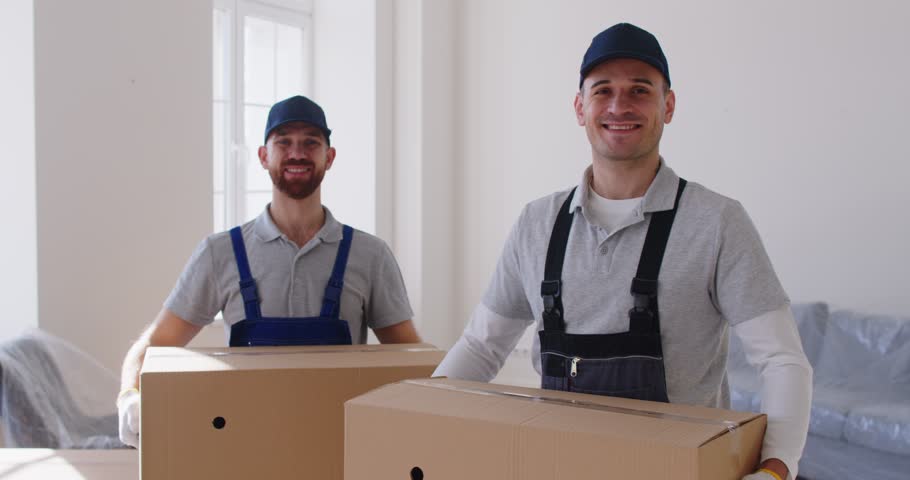 Portrait of two professional workers looking at camera, carrying cardboard boxes, helping with moving to new apartment. Employees of moving company delivering and unloading boxes during relocation.