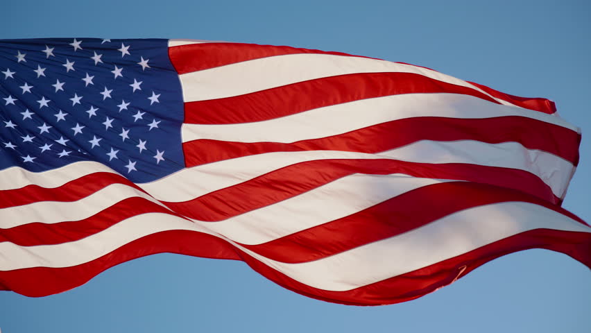 Close-up of the American flag waving in the wind against a clear blue sky. Captured in 120fps slow motion, highlighting the elegant motion of the fabric and vibrant red, white, and blue colors.