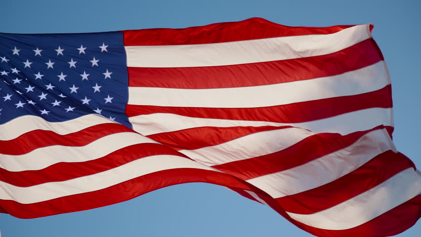 Close-up of the American flag waving in the wind against a clear blue sky. Captured in 120fps slow motion, highlighting the elegant motion of the fabric and vibrant red, white, and blue colors.