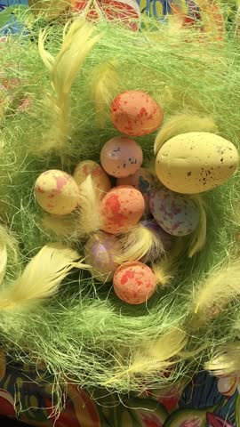 A festive Easter nest with green fiber grass, colorful speckled eggs, and yellow feathers, rotating clockwise (around the clock) on the tropical floral background; a vertical slowmotion video.