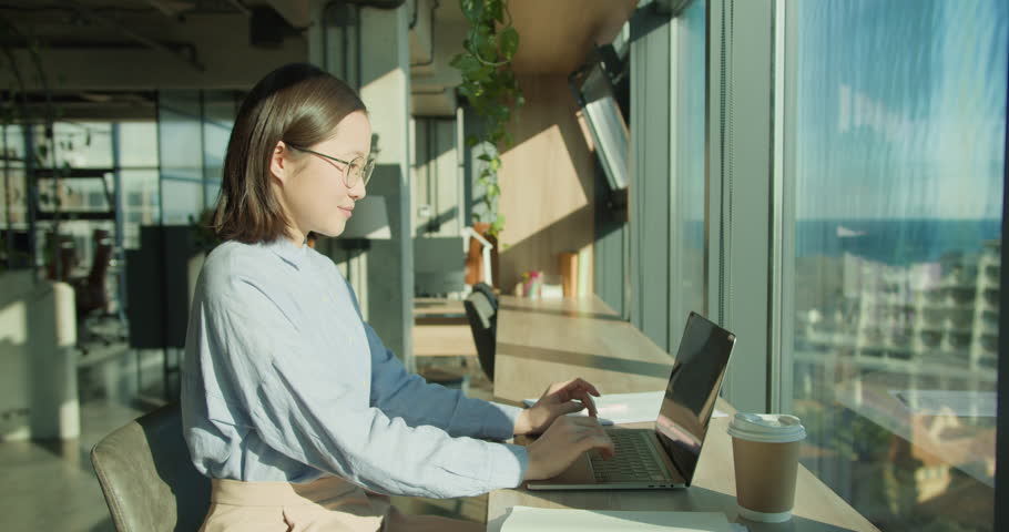 A young woman in a bright modern office, enjoying a calm moment at her desk with a laptop, notebook, and coffee, surrounded by greenery and natural sunlight