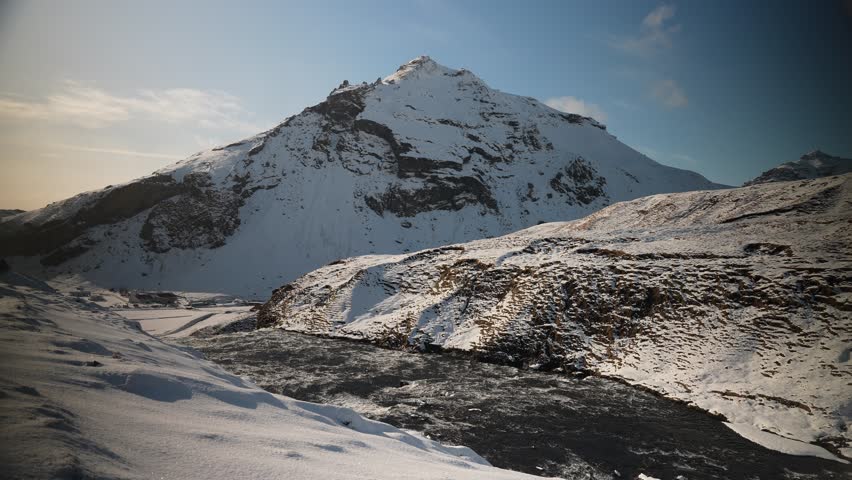 Video of a river flowing over a snow capped mountain with water rushing over rocks in the foreground and a tall snow capped mountain under blue skies in the background. 