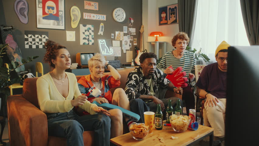 Group of young diverse friends sitting on couch in cozy living room with snacks and drinks on table, watching sports game on TV and getting disappointed