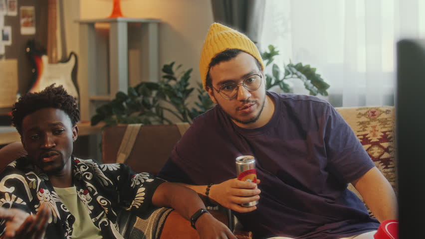 Group of joyful diverse friends sitting in living room, drinking beer, chatting and laughing, having fun while watching sports competition on TV together at home