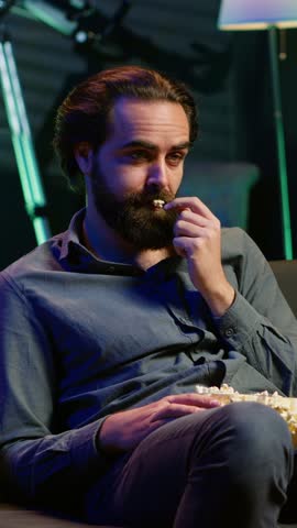 Vertical video Exhausted man watching shows on TV late at night begore going to sleep and eating popcorn. Sleepy man in pajamas eating snack on couch, consuming mindless entertainment, camera A