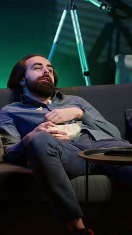 Vertical video Bored man at home entertaining himself by throwing popcorn in air and catching it with mouth, wasting time. Person slacking off, eating salty snacks and lounging on couch watching TV