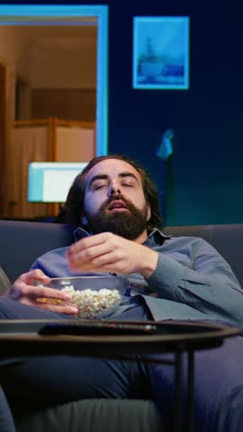 Vertical video Bored man at home entertaining himself by throwing popcorn in air and catching it with mouth, wasting time. Person slacking off, eating salty snacks and lounging on couch watching TV