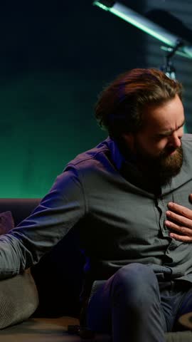 Vertical video Man in apartment suffering heart attack while watching movie on TV, living troublesome sedentary lifestyle. Sick person feeling pain in chest, trembling on couch, fighting for his life