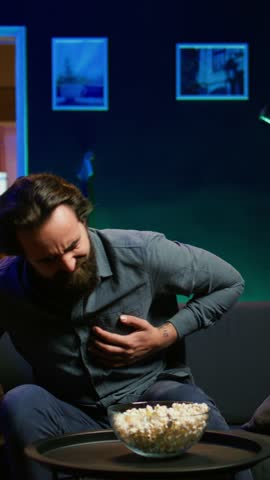 Vertical video Man in apartment suffering heart attack while watching movie on TV, living troublesome sedentary lifestyle. Sick person feeling pain in chest, trembling on couch, fighting for his life