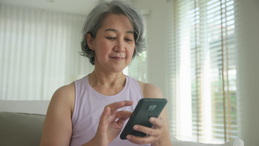 Asia adult people middle age happy relax old woman using mobile ask AI chatGPT chat bot look health care app sit at home sofa elder aging asian aged female. Online social media smart pay order system.