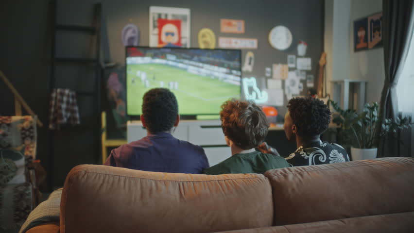 Three young men sitting on couch, raising hands and cheering while watching soccer match on TV in stylish living room with modern decor. Rear view