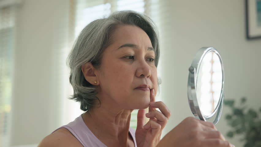 Asia adult people middle age old woman look at mirror dry skin face hand touch neck cheek stress sad at home elder health care aging beauty issue. Asian female aged eye botox filler plastic surgery.