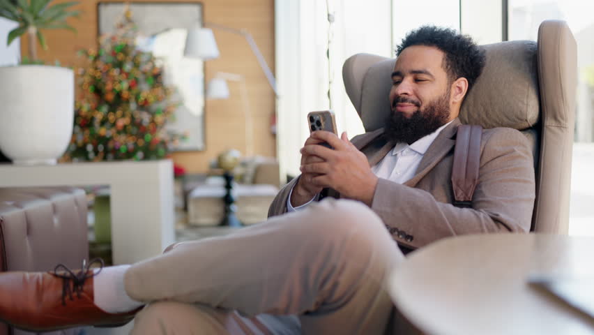 Stylish African American man relaxes in a cozy armchair, engaged with his smartphone, enjoying the bright space around him, showcasing modern lifestyle and leisurely moments of contemporary living