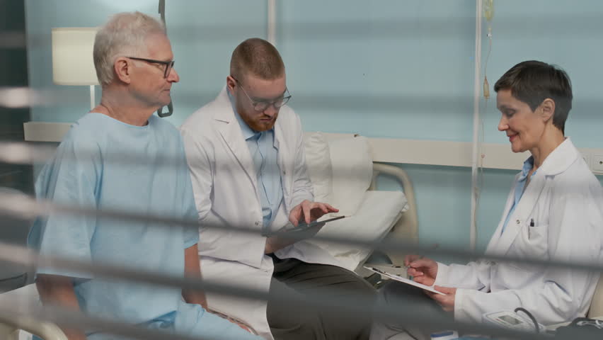 Medium long shot of two doctors in white coats using digital tablet while discussing treatment plan with male senior patient in hospital ward