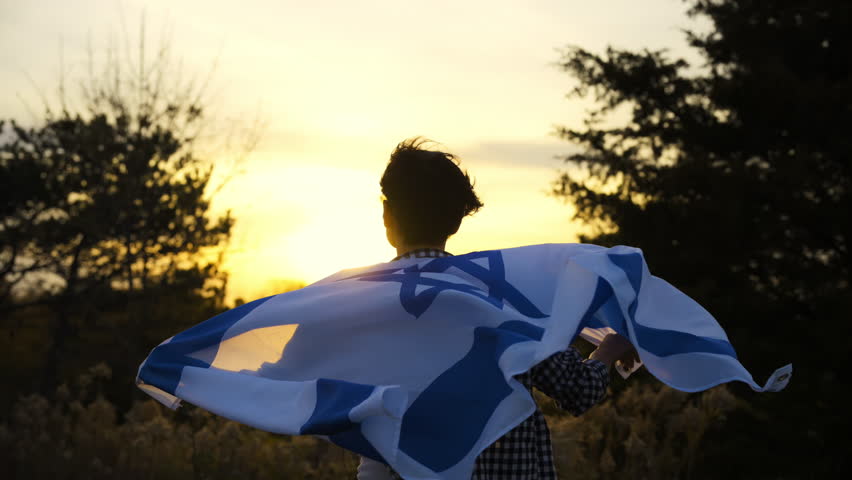 A woman standing with the Israel flag against a strong wind on the background of clear sky at sunset.