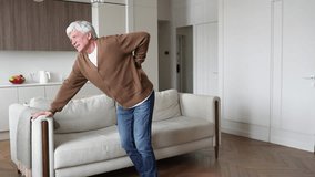 Agonized weak tormented elderly man with backache painfully attempting walk, leaning on sofa. Pinched nerves, herniated disc, nerve compression, chronic suffering, spinal disorder, physical torment - Powered by Shutterstock - Get 15% off with code: PIKWIZARD15