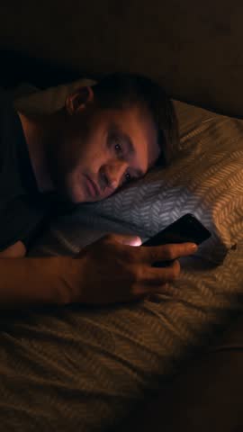 Exhausted man in bed at a very late hour quietly scrolls phone with tired eyes, reveals insomnia, anxiety, and deep digital dependency