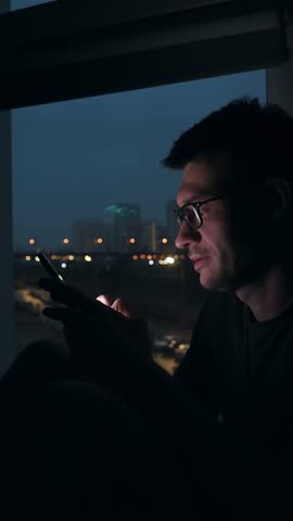 Smiling young man in glasses typing message on smartphone at night, city lights glow behind window, pleasant texting in urban apartment.