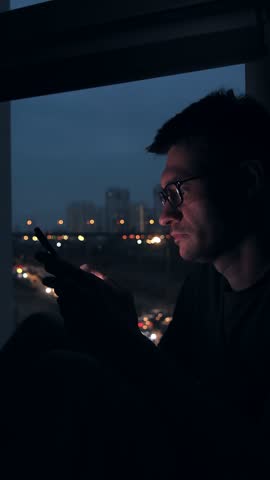 Focused young man in glasses scrolling smartphone screen at night by window, city lights reflecting, browsing content in quiet apartment.