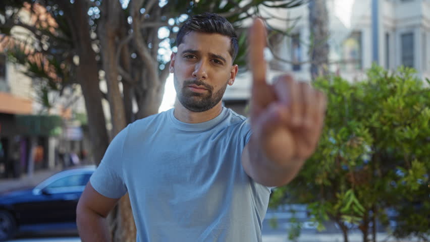 Young man in a city park says no with his finger, highlighting his confident expression in an urban outdoor setting with trees and streets in the background.