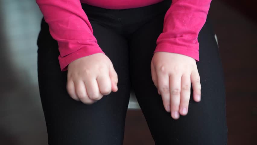 Girl places hands on knees, moves fingers. She spreads and closes her fingers repeatedly. Fingers stretched wide. Child wears long-sleeve pink shirt, black pants. Close-up of hand movement, posture