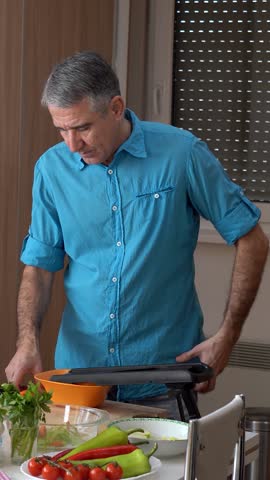 Casual Middle-aged Man with Gray Hair, Slices Carrots at Home on a Table full of Various Vegetables. The video is perfect for promoting healthy food, food tutorials and lifestyle of middle-aged men.