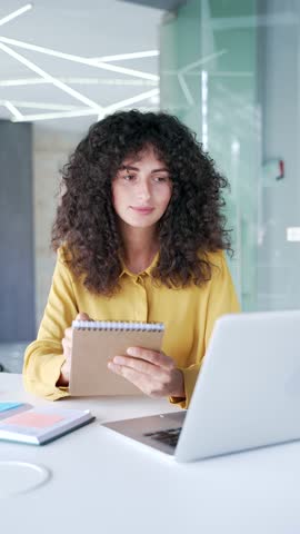 Young adult female employee watching video call training notes in notebook looking at laptop screen sitting in business office. Businesswoman is studying online, listening remotely to course. Vertical