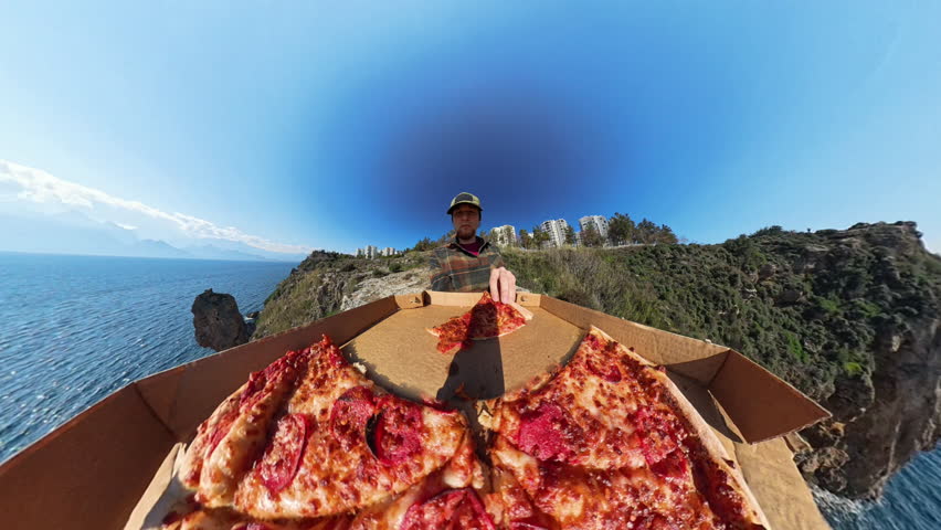 Man Standing on a Cliff Eating Pizza with a Large Open Pizza Box in the Foreground, Captured with a Wide-Angle Effect and a Panoramic Ocean View in the Background