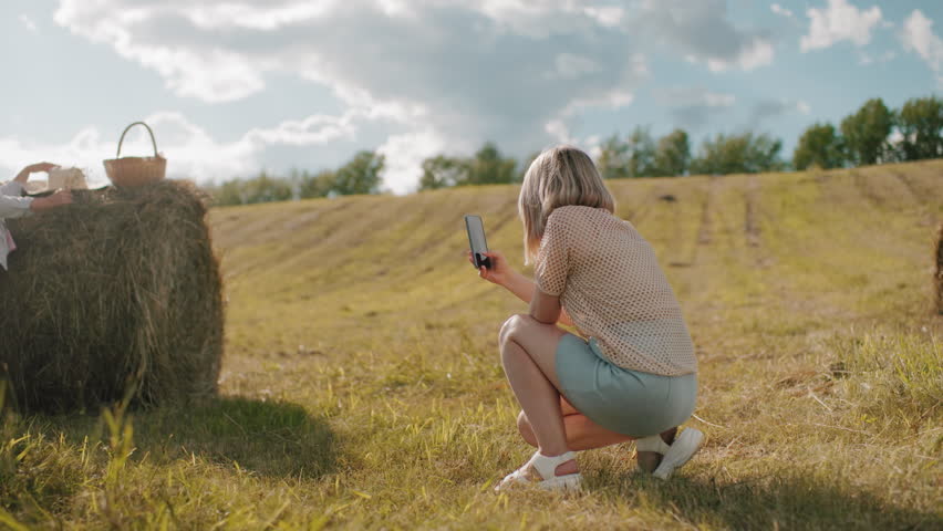 Rear view of lady squatting taking photos of her sister in vast farmland with blurred background, warm countryside scene with hay bale and golden sunlight