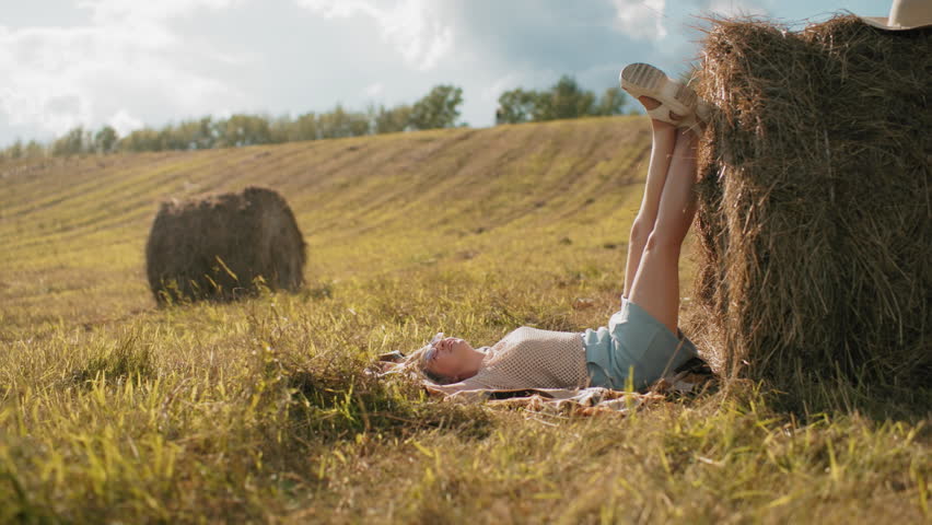 Lady squatting taking sister photos outdoors as she places legs on hay in vast farmland, warm countryside setting with golden fields, rustic atmosphere, and sunlight casting soft shadows