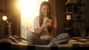 Happy young woman texting with friends on smartphone while sitting on bed in cozy bedroom at sunset - Powered by Shutterstock - Get 15% off with code: PIKWIZARD15