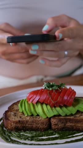 Vertical screen, woman using smartphone taking pictures of her avocado toast with tomatoes and pesto sauce on whole wheat bread, ready to share on social media