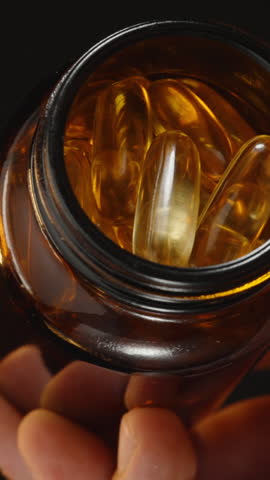 Vertical video. Hand Pouring Omega 3 Capsules from a Brown Glass Bottle into an Open Palm, Capturing the Motion Against a Dark Background