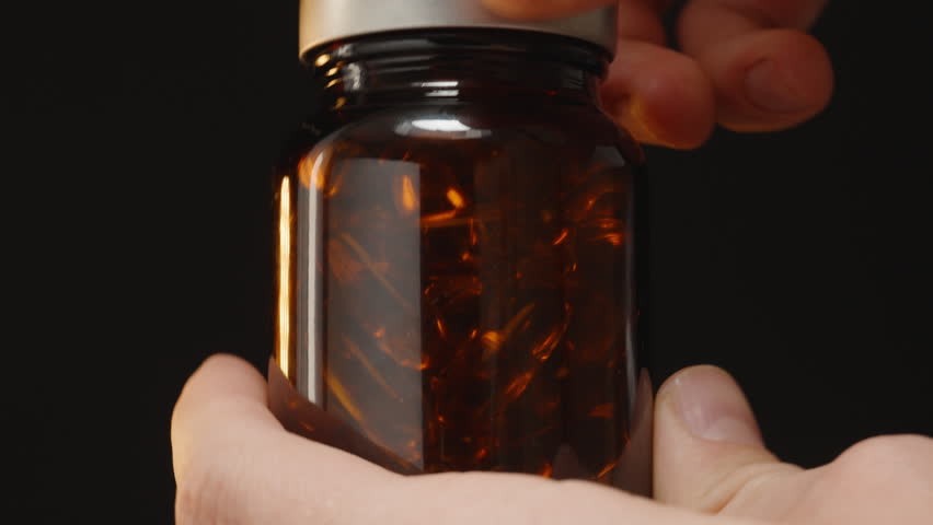 Hand Dropping an Omega 3 Capsule into a Brown Glass Bottle with an Open Neck, Capturing the Motion Against a Dark Background
