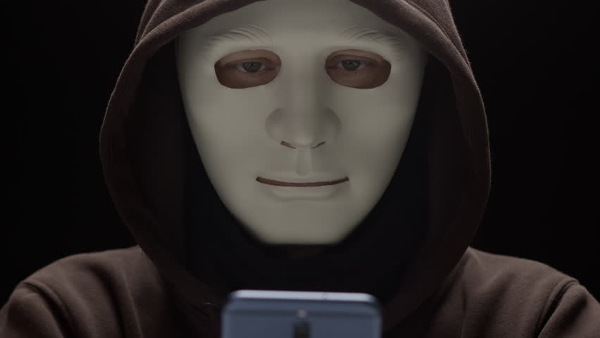 Masked criminal uses phone to commit fraud. Concept of hacking and cybersecurity via smartphone.