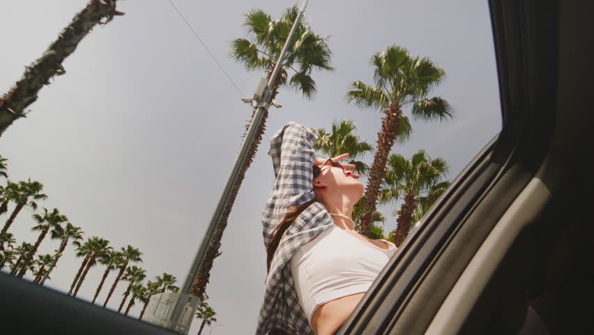 Excited woman enjoys the palm trees of seaside road. Girl leans out of the window of a rental car on a coastal highway on a sunny day. Hair in wind. A tropical vacation on the beach of exotic islands