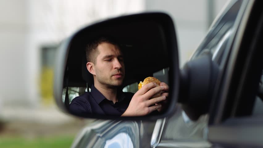 Young man inside car. A view in side mirror of guy in shirt eating burger. Breakfast or lunch in car. Eating and enjoying meal. Slow motion.
