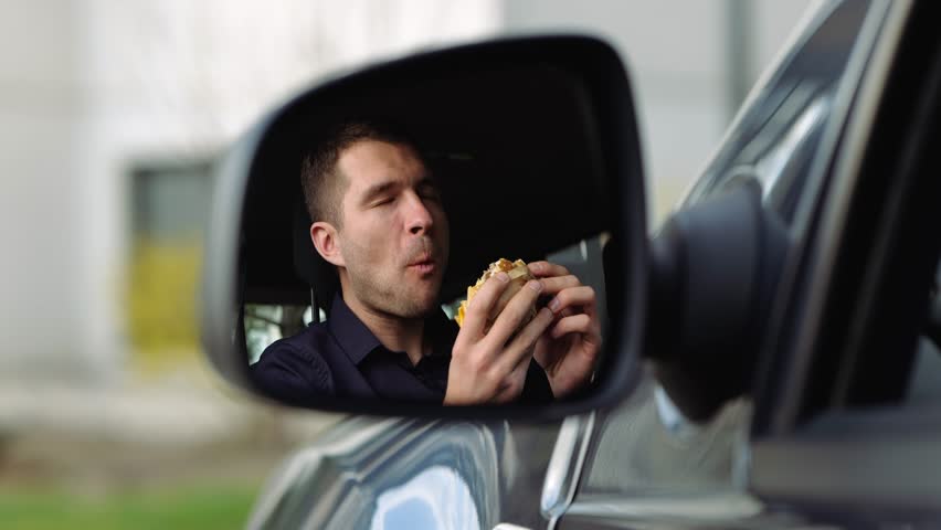 Young man inside car. A view in side mirror of guy in shirt eating burger. Breakfast or lunch in car. Eating and enjoying meal. Slow motion.