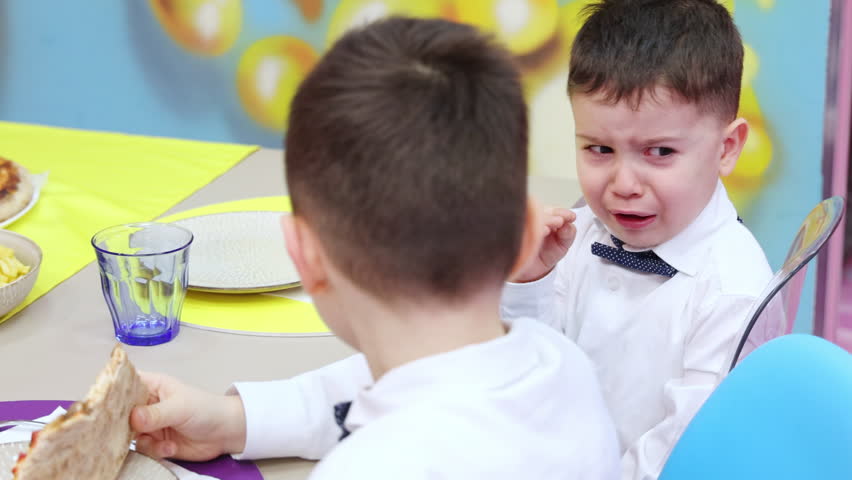 kids sitting at table on birthday party. one boy is eating pizza and another is crying,makins whims. colorful wall in background.child in shirt with bow on neck.